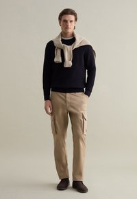 Marineblauer Strickpullover, beiger Cargo-Hose mit Seitentaschen und braune Ankle Boots. Pullover über die Schultern drapiert. Schlichtes, funktionales Design.
