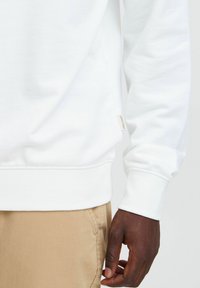Felpa bianca con polsini e orlo a costine, materiale in cotone, dettaglio dell'etichetta laterale. Pantaloni beige visibili nella parte inferiore dell'immagine, mostrano la tasca.