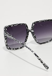 Black and white sunglasses with a bold, patterned frame and gradient gray lenses; oversized square shape and slim, patterned arms.