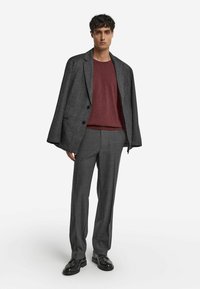 Grau strukturierter Blazer, burgunderfarbener Strickpullover, graue maßgeschneiderte Hose und schwarze Lederschuhe. Modell steht aufrecht und blickt nach vorne.