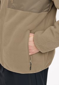 Hand inserting into beige fleece jacket side pocket with zipper, showing soft textured sleeve and black pants below.