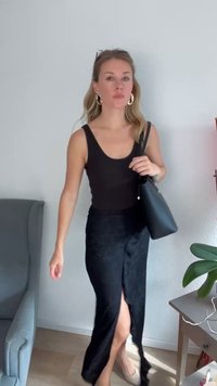 Top preto sem mangas e uma saia preta estampada com uma fenda. Ela carrega uma bolsa preta e usa brincos, de pé contra uma parede clara.