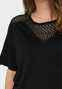 Svart t-shirt med kort ärm, utformad med en spetsinsättning vid halsringningen. Den har en slät yta och en lätt stretch i tyget.