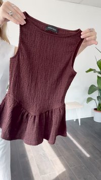 Person holding a sleeveless textured dark red peplum top with an "Anna Field" label in a bright room with plant and bench.