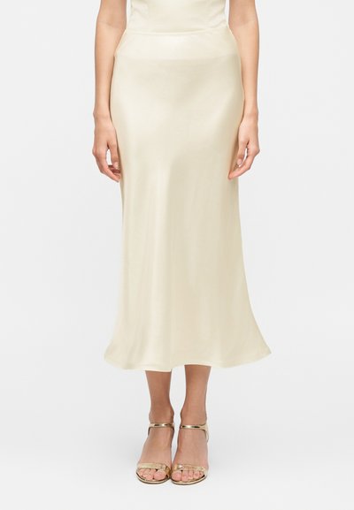 Femme portant une jupe en satin couleur crème jusqu'aux chevilles et des sandales à talons hauts dorées à bout ouvert, debout les bras détendus le long du corps.
