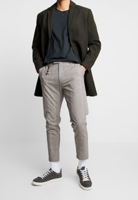 Olive green long blazer, navy t-shirt, gray plaid trousers, white socks, and dark sneakers with white soles. Polished look with layered textures.