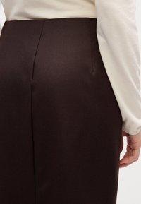 Gros plan d'une personne portant une jupe marron foncé et un haut crème à manches longues, la main droite reposant le long du corps.