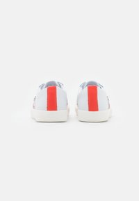 Ett par vita sneakers med klara röda vertikala ränder på hälarna, visade mot en helt vit bakgrund, sedd bakifrån.