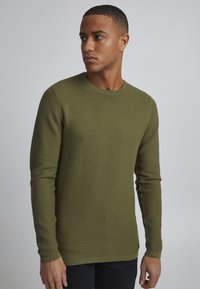 Pulover lung cu mâneci lungi, de culoare verde măslinie, cu o textură cu nervuri și guler rotund. Fabricat din material tricotat, cu un design ajustat și detalii de coasere minime.