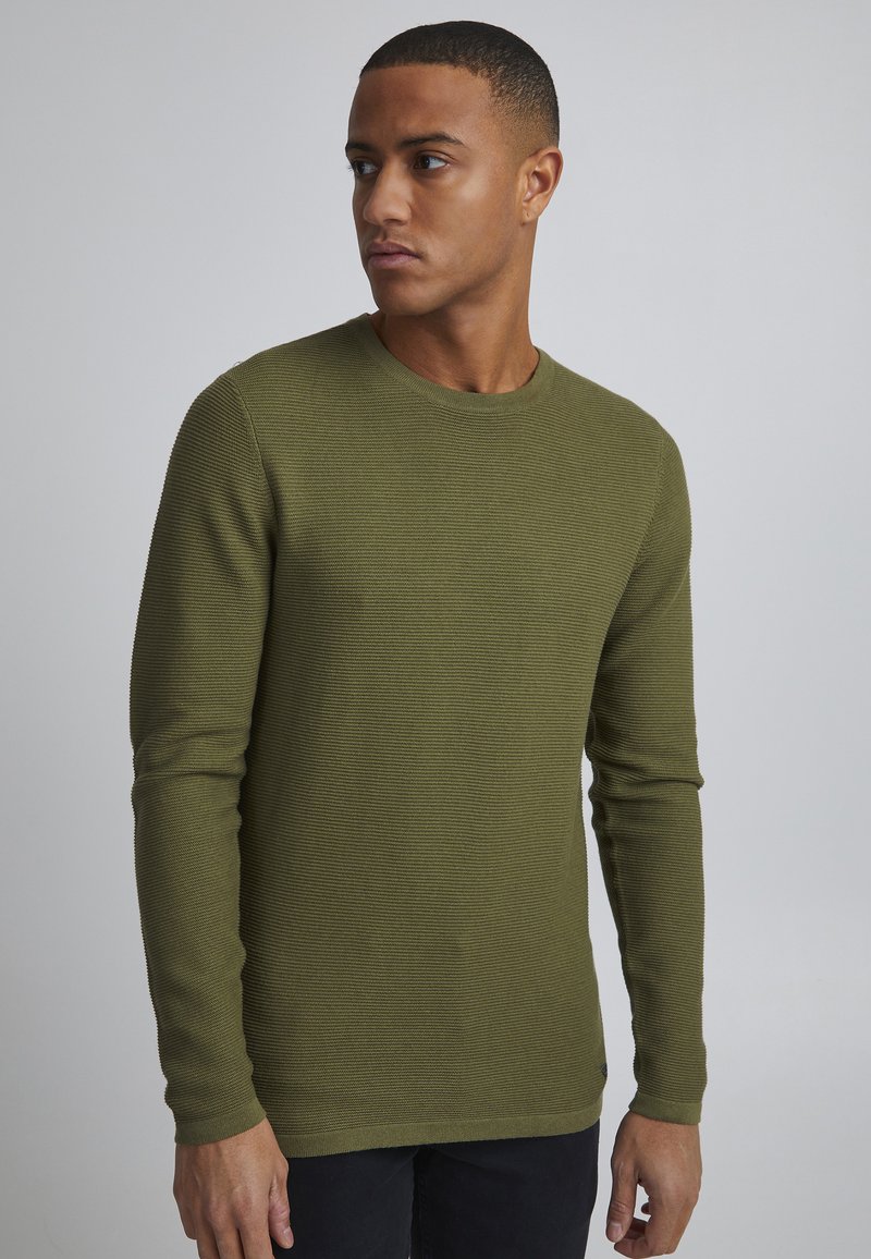 Pulover lung cu mâneci lungi, de culoare verde măslinie, cu o textură cu nervuri și guler rotund. Fabricat din material tricotat, cu un design ajustat și detalii de coasere minime.
