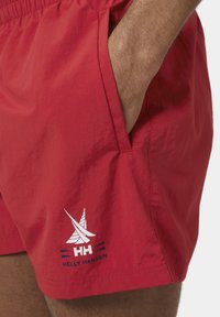 Pantaloni da nuoto rossi con elastico in vita, tasche laterali e un logo "Helly Hansen" bianco che presenta una barca a vela e accenti blu. Texture liscia.