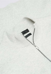 Pull gris clair tricoté avec une courte fermeture éclair partiellement ouverte au niveau du col, montrant deux étiquettes noires à l'intérieur du col.