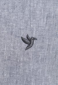 Tejido gris con un acabado texturizado que presenta un pequeño colibrí negro bordado en el lado izquierdo. Diseño simple y detallado.