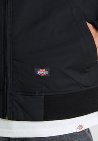 Čierna kapucňová bunda vyrobená z hladkej látky, s predným zipsom, rebrovanými manžetami a vyšitým logom Dickies na prsiach.