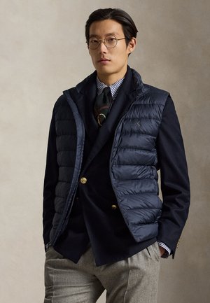 Homme portant des lunettes rondes, une doudoune bleu marine sur un blazer noir, une chemise rayée, une cravate à motifs et un pantalon gris sur un fond uni.