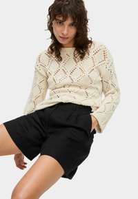 Crème gebreide trui met diamantvormige uitsparingen, gecombineerd met zwarte high-waisted shorts. De trui heeft lange mouwen en een geribbelde halslijn.