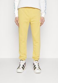 Gula mjukisbyxor med elastiska muddar och dragsko i midjan; har en liten logotyp på vänstra låret. Bärs med svartvita sneakers.