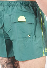 Pantaloni da bagno verdi realizzati in tessuto leggero, dotati di una tasca laterale con logo giallo, cuciture a contrasto e una vita elasticizzata.