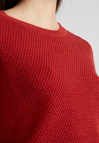 Pull rouge en tricot avec un tissage texturé, présentant un col rond classique et un ourlet côtelé. Vue rapprochée du tissu et des détails du design.