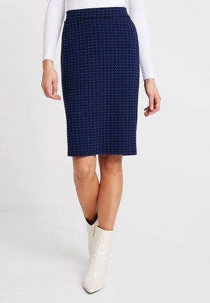 Marineblauwe pied-de-poule kokerrok, knieachtig, soepele textuur, hoge taille, gecombineerd met een witte aansluitende top met lange mouwen en crème enkellaarsjes.