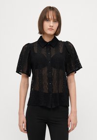 Blusa nera in pizzo trasparente con maniche a palloncino corte, caratterizzata da una chiusura con bottoni sul davanti, colletto e intricati motivi floreali su tutta la superficie.