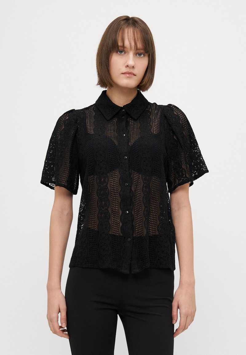 Blusa nera in pizzo trasparente con maniche a palloncino corte, caratterizzata da una chiusura con bottoni sul davanti, colletto e intricati motivi floreali su tutta la superficie.
