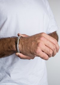 Pintas sidabrinis apyrankė su cilindriniu metaliniu užsegimu, gulinčiu ant riešo. Fonas yra neryškus ir baltas, išryškinantis apyrankę.