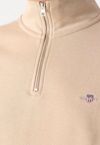 Sudadera cremosa con cremallera y alto cuello, que cuenta con una cremallera plateada y un pequeño logo bordado en el pecho. Tejido liso y texturizado.