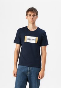 Marineblå bomulls T-skjorte med en rektangulær hvit og gull logo etikett med teksten "JACK & JONES" over brystet. Korte ermer, rund hals.