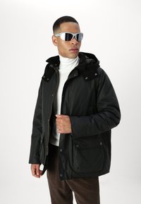 Μαύρο αδιάβροχο τζάκετ με φερμουάρ και κουμπί, που διαθέτει γιακά επενδεδυμένο με σουέτ και δύο μπροστινές τσέπες. Φορεμένο πάνω από λευκό πουλόβερ ψηλού λαιμού.