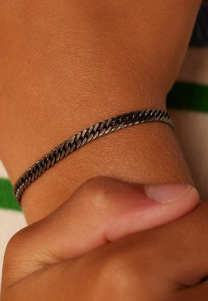 Bracelet chaîne en argent au poignet, une main tenant doucement le poignet sur un fond flou vert et blanc.