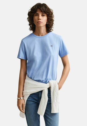 Frau mit lockigem Haar trägt hellblaues T-Shirt, weißen Pullover um die Taille gebunden, blaue Jeans und silbernes Armband, steht vor einfarbigem Hintergrund.