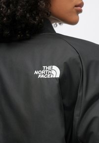 Czarna kurtka bomber z prążkowanym kołnierzem i białym haftowanym logo, gładka w dotyku z lekko błyszczącym wykończeniem.