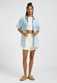 Camisa de mezclilla azul clara de manga corta, con dos bolsillos frontales, usada abierta sobre un crop top blanco y un pantalón corto blanco, combinada con zapatos sin hebilla color marrón claro.