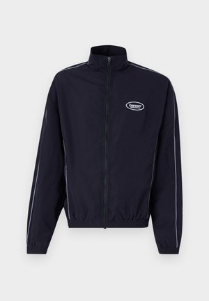 Veste coupe-vent marine avec un col montant, zip complet, taille élastique et rayures blanches contrastées le long des manches. Présente un logo.