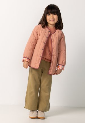Lichtroze gewatteerde jas met drukknoopsluitingen, paarse afwerking en kleurrijke met bloemenpatroon versierde mouwen. Gecombineerd met beige wijde broek en tankleurige schoenen.