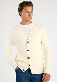 Krämfärgad ribbad cardigan med V-ringning, fem svarta knappar fram, och långa ärmar, bärs över en vit T-shirt och blå jeans.