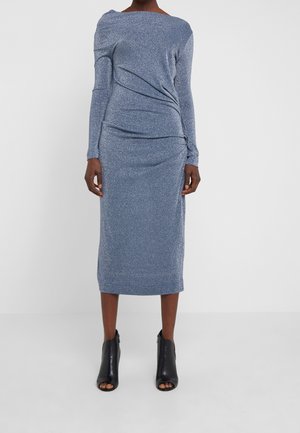Femme portant une robe midi bleue étincelante à manches longues avec des détails drapés, associée à des bottines noires à bout ouvert sur un fond uni.