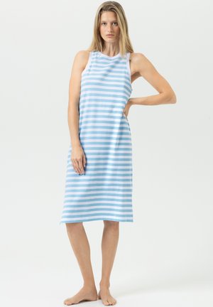 Jeune femme debout, pieds nus, portant une robe sans manches longueur genou avec des rayures horizontales bleu clair et blanches sur un fond uni.