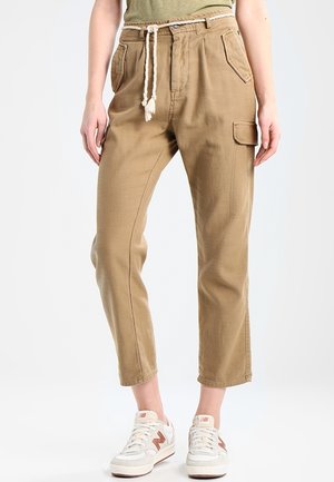 Kvinde iført beige cropped cargobukser med forlommer og reb-bælte, kombineret med hvide sneakers på en ensfarvet baggrund.