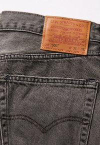 Grå denimjeans med en brun läderetikett märkt "Levi Strauss & Co." och "Lot 501." Har klassiska detaljer med stygn på bakfickan.