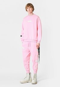 Sudadera rosa y pantalones jogger a juego hechos de una mezcla de algodón. Presenta una etiqueta en el pecho y detalles en contraste en los pantalones. Botas blancas.