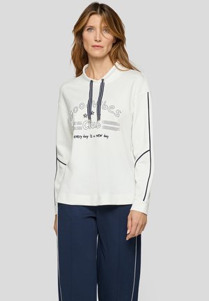 Frau trägt weißen Kapuzenpullover mit langen Ärmeln und Aufschrift "Good Vibes Club" sowie marineblaue Kordeln, kombiniert mit marineblauen Hosen mit weißen Seitenstreifen.