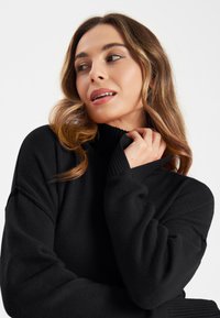 Zwarte coltrui van een zachte breimateriaal met geribbelde kraag en manchetten. Het heeft een relaxte pasvorm en een design met verlaagde schouders.