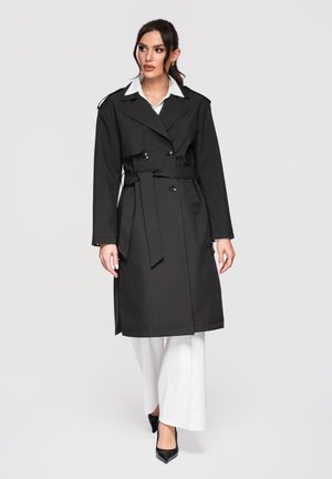 Donna con capelli scuri indossa un trench nero con cintura sopra pantaloni bianchi a gamba larga e tacchi neri, in piedi davanti a uno sfondo chiaro.