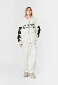 Sudadera blanca con cremallera y texto gráfico negro en las mangas, combinada con pantalones blancos a juego. Tejido suave, corte relajado y cordones ajustables.