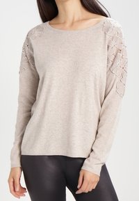 Beige Pullover mit langen Ärmeln aus weichem Strickmaterial, mit Spitzendetails in Blumenmuster an den Schultern und einer lockeren Passform.