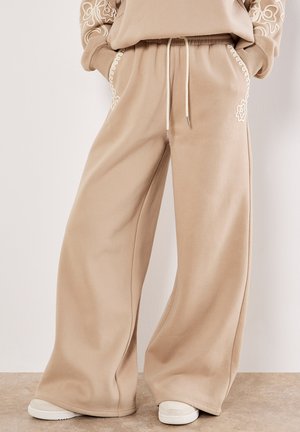 Pantalons de survêtement larges beige en tissu doux, dotés d'un motif floral brodé le long des poches et d'une taille à cordon.