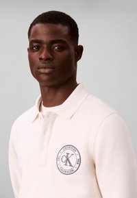Νέος άνδρας φοράει λευκό πόλο με λογότυπο Calvin Klein στο αριστερό στήθος, κοιτάζοντας προς τα εμπρός με ουδέτερη έκφραση.