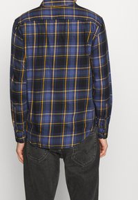 Chemise à carreaux bleu et noir avec des accents jaunes, des poignets à boutons et un ourlet arrondi. Le tissu semble doux avec un motif à carreaux classique.